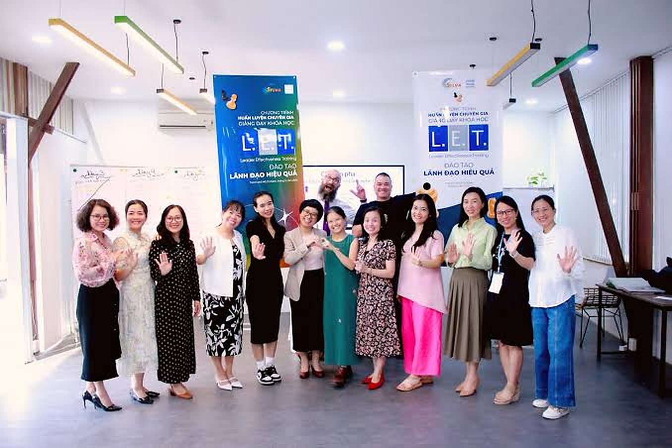 Mười bốn người đứng thành hàng, vẫy tay chào trước hai băng rôn chương trình huấn luyện giảng viên L.E.T.